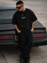 A man wearing a black t-shirt with 'VAKO' printed in white on the chest, black sweatpants, and sunglasses, standing in front of a black car.