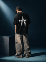 A person stands with their back to the camera wearing a black t-shirt with a large distressed white star graphic on the back, paired with baggy faded gray cargo pants and a black baseball cap, set against a dark blue studio background with a spotlight.