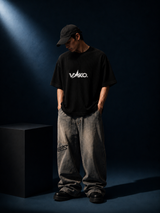 A person wearing a black VAKO. t-shirt, gray washed jeans, and a black cap, standing in a dark studio setting with dramatic spotlighting.